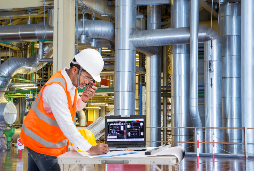 Engineer making notes on his notebook with a laptop screen on the side in a powerhouse/