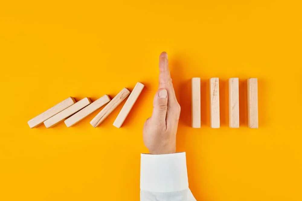 A hand stopping a line of falling dominoes, preventing the rest from toppling, on an orange background.