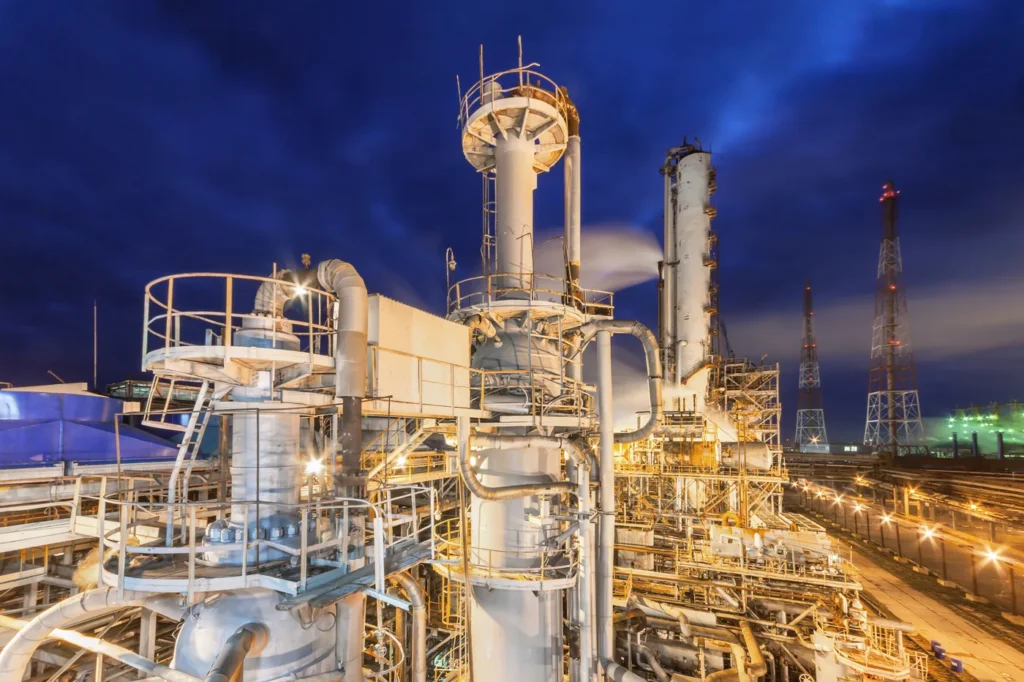 Large petrochemical refinery lit at dusk, with distillation columns, flare stacks and pipework against a deep blue sky.