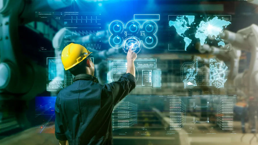 Worker in a yellow hard hat touching a large translucent interface of dashboards, gauges and a world map, with machinery behind.