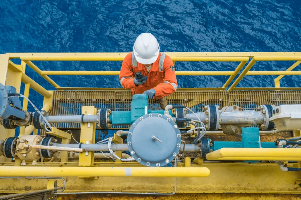 Offshore worker in orange coveralls and a hard hat servicing a valve assembly on a platform walkway above the sea.