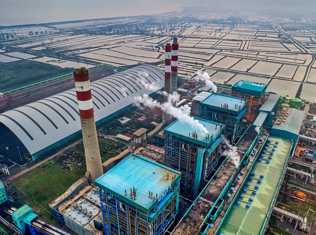 Aerial view of an industrial plant with tall striped chimneys emitting steam, beside large storage buildings and open land.