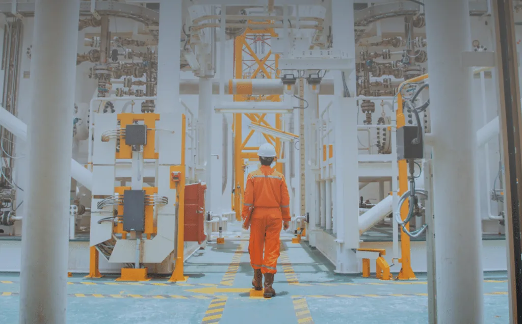 Worker in orange coveralls and a hard hat walking through an industrial plant between rows of equipment and pipework.