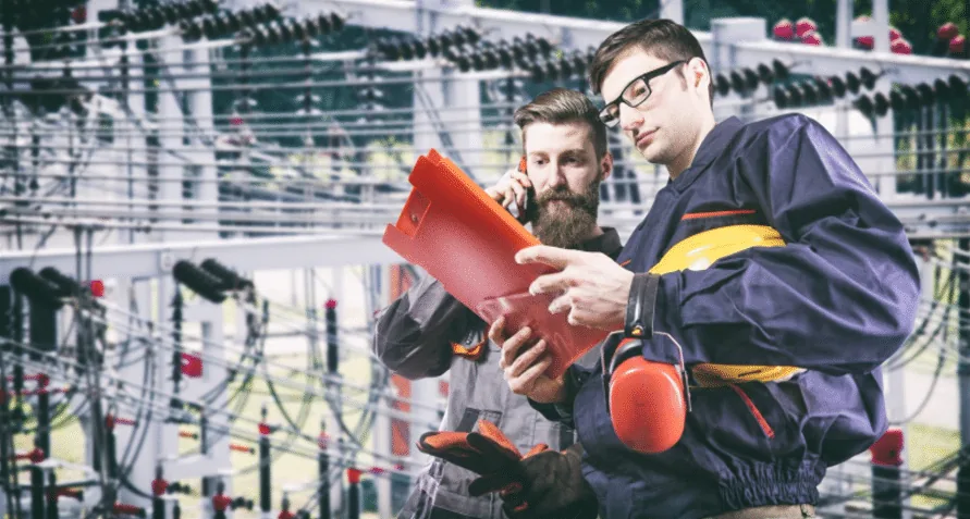 Two technicians in workwear consulting a folder beside high-voltage substation equipment, one on the phone.