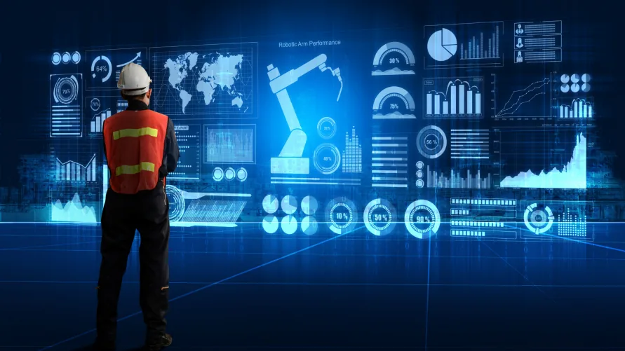 Worker in a hard hat and high-visibility vest standing before a large holographic display of dashboards, charts and a robotic arm.