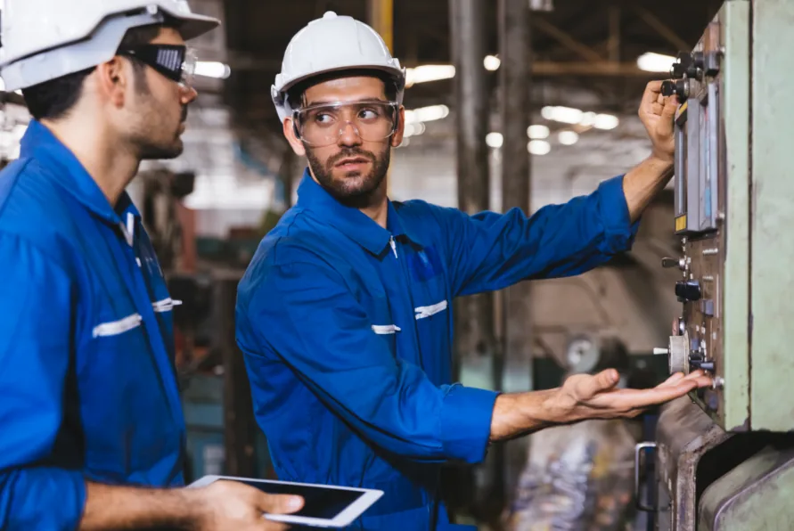 Two workers in blue coveralls and hard hats inspecting a control panel in a plant, one holding a tablet.