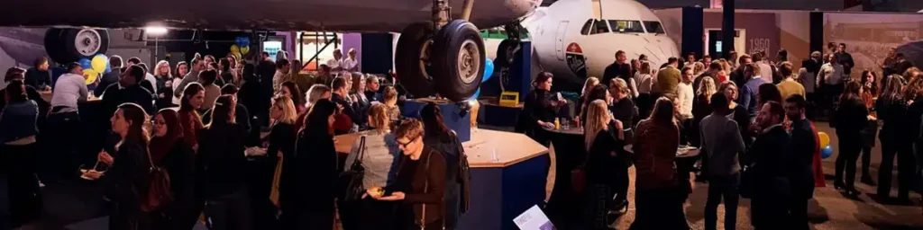 Busy networking event in an aircraft hangar, with attendees standing among exhibition stands beside a parked aeroplane.