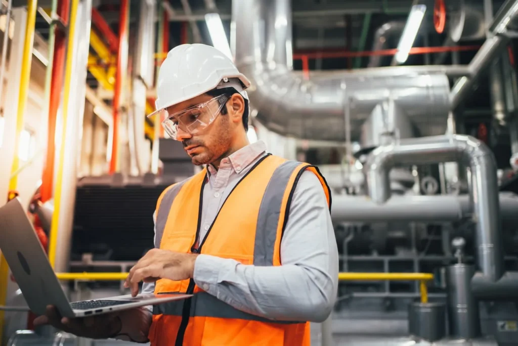 Engineer in a hard hat, safety glasses and high-visibility vest using a laptop in front of industrial pipework.
