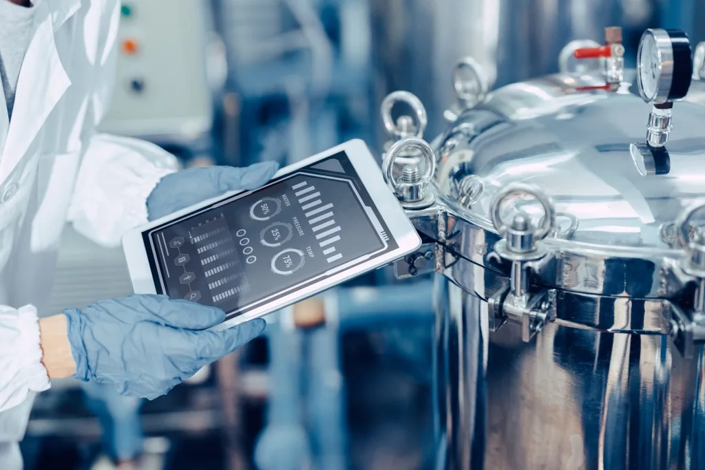 Gloved technician holding a tablet showing a control interface beside polished stainless steel process vessels.