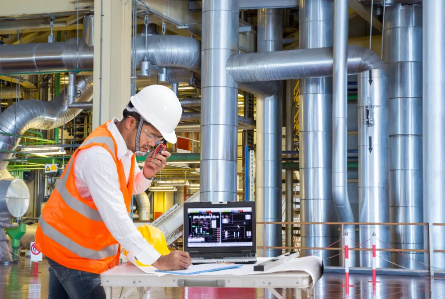Engineer in a hard hat and high-visibility vest making notes at a laptop set up beside large insulated pipework in a plant.