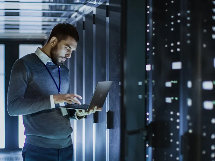 IT engineer working on a laptop while standing in a data centre aisle between server racks.