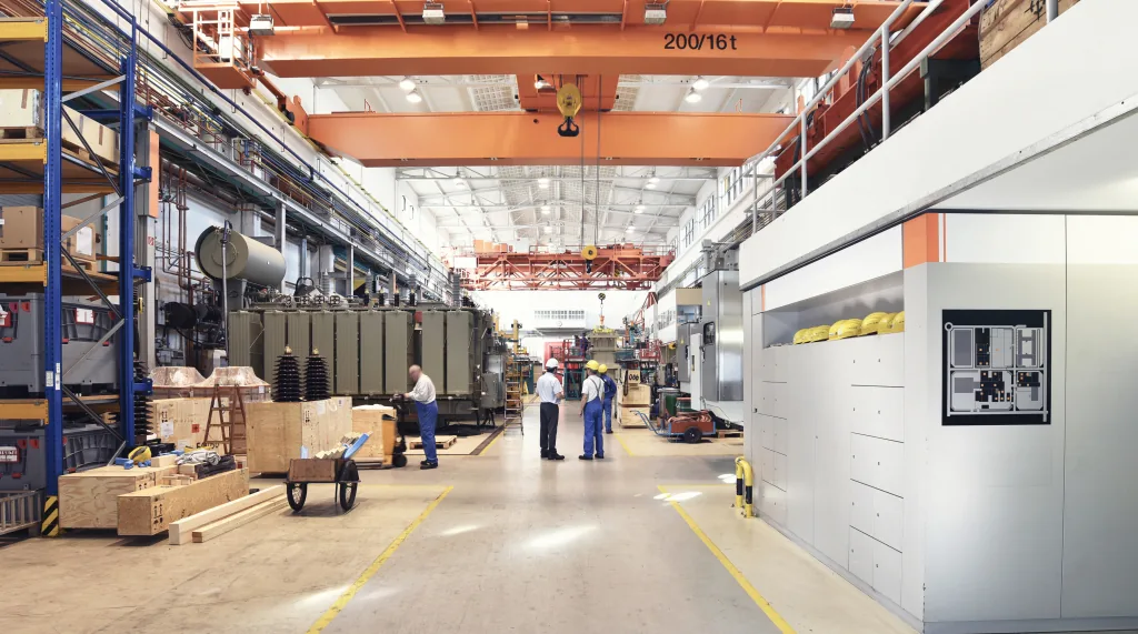 Interior of a manufacturing hall with an overhead crane rated 200 by 16 tonnes, stacked components, workers in the aisle and a large enclosed machine to the right.