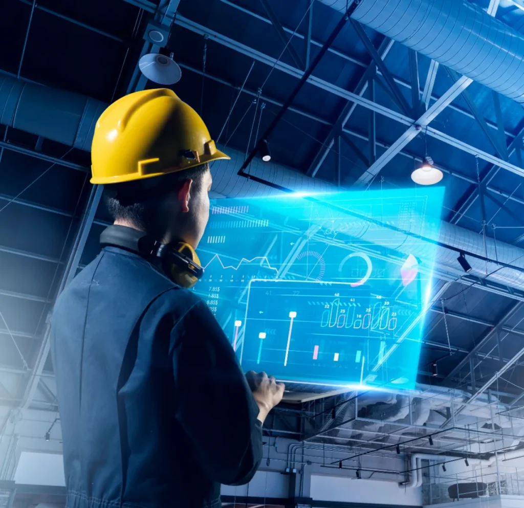 Worker in a yellow hard hat and ear defenders viewing a glowing holographic dashboard inside an industrial hall.
