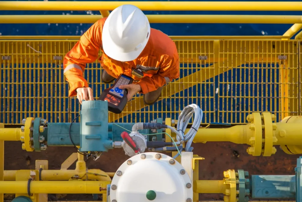 Offshore worker in orange coveralls and a white hard hat crouching over yellow pipework, using a handheld device on an instrument.