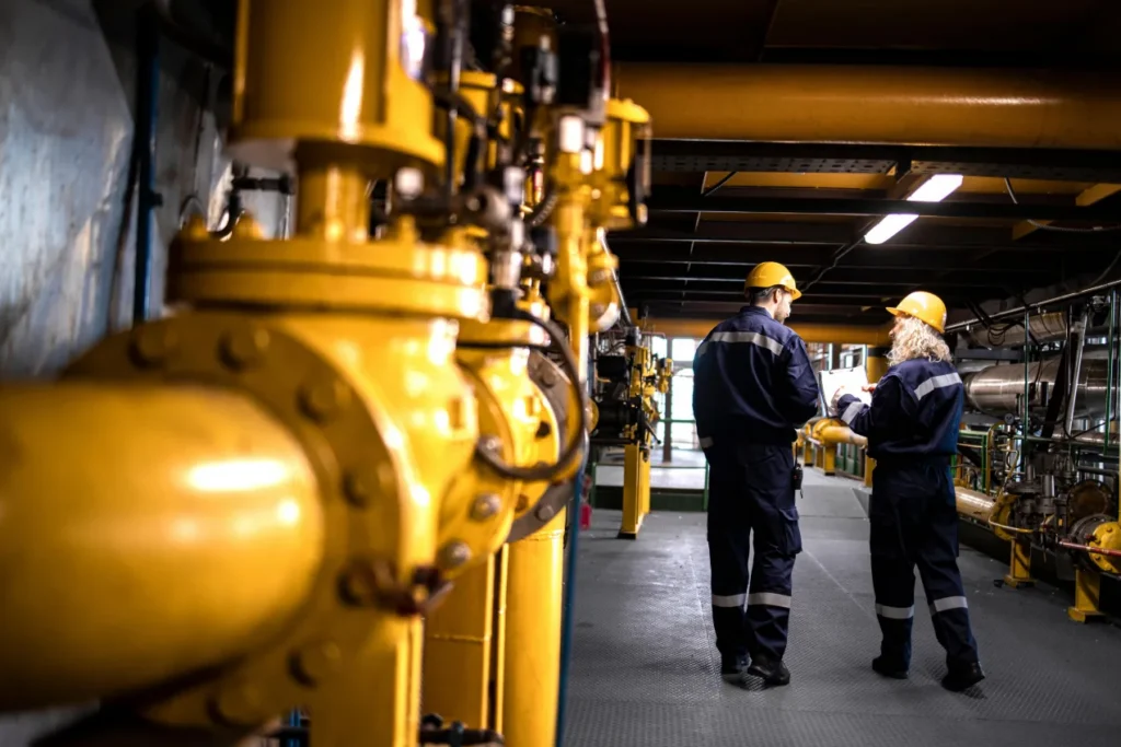 Two workers in coveralls and hard hats walking through a plant corridor lined with yellow pipework and valves.
