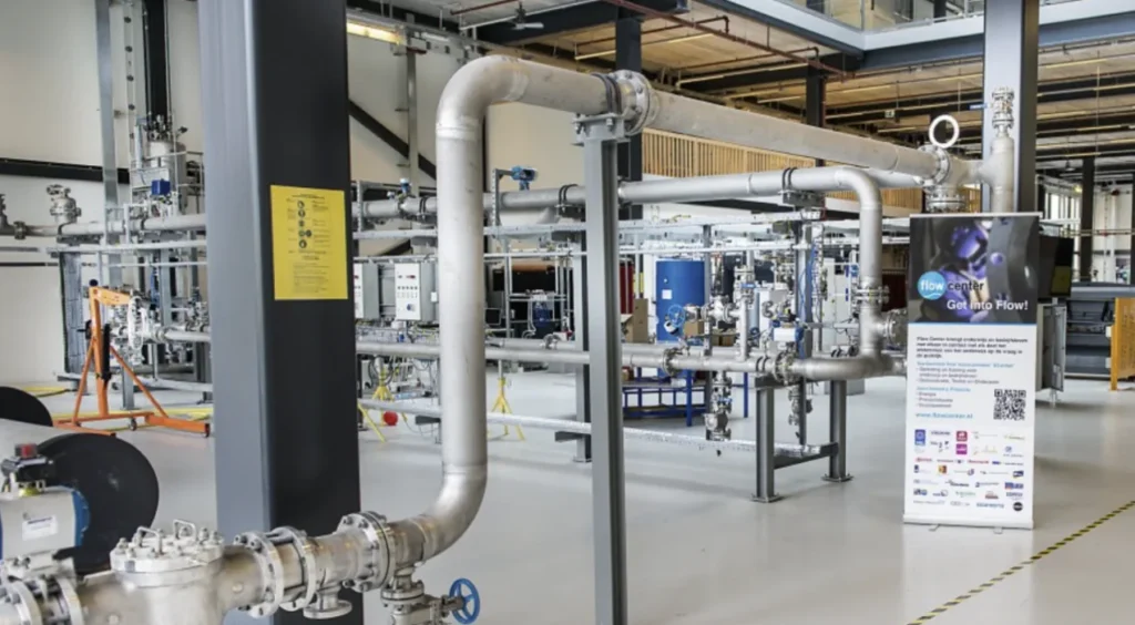 Demonstrator flow rig in a workshop, with stainless steel pipework, valves and instrumentation, and an information stand beside it.