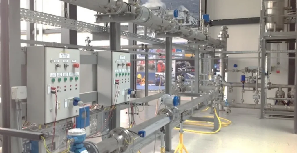 Pilot plant skid with stainless steel pipework, instrumentation and control cabinets in a laboratory hall.
