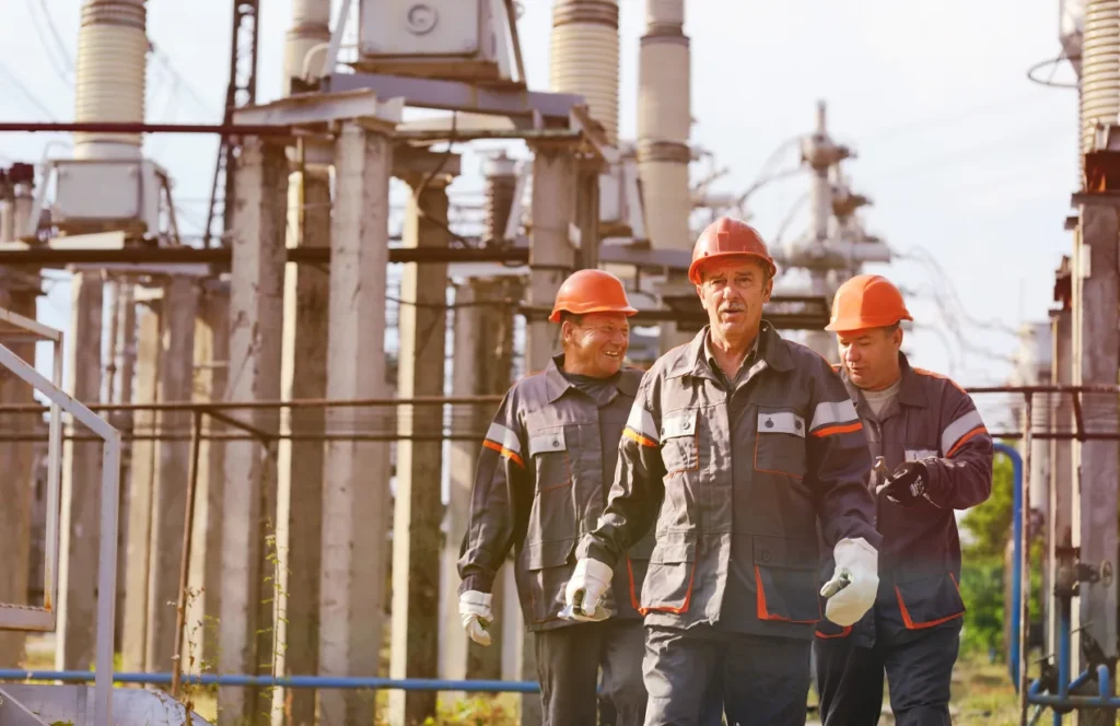 Three workers in hard hats and overalls talking beside high-voltage substation equipment, one holding a tablet.