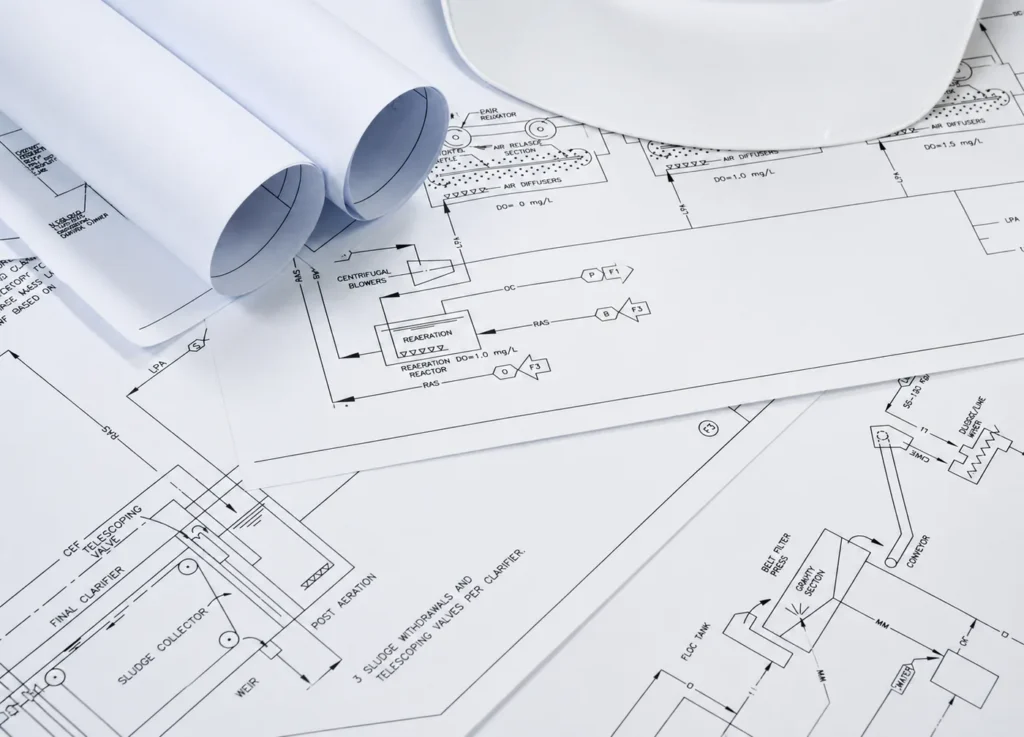 Rolled and flat engineering drawings of a process plant spread across a desk, showing piping and instrumentation schematics for clarifiers, blowers and valves.