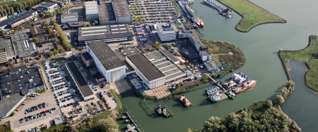 Aerial view of a shipyard and industrial site beside a river, with workshops, parked vehicles and vessels at the quayside.