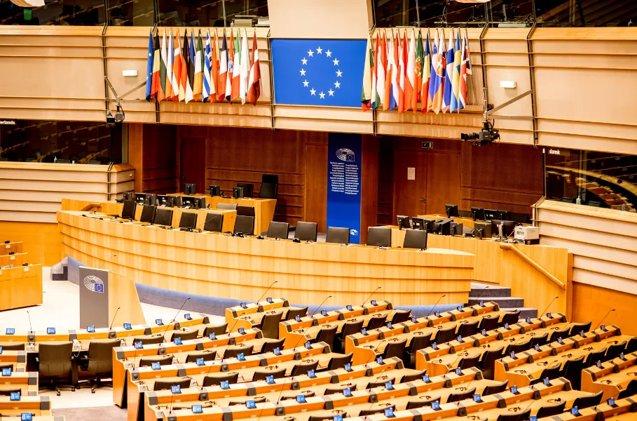 Empty debating chamber of the European Parliament, with member state flags and the EU flag behind the podium.