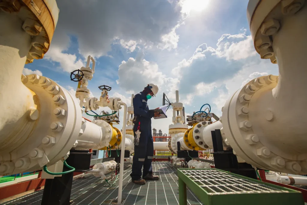 Worker with a tablet standing between large wellhead valves on an offshore platform, seen from below against a bright sky.