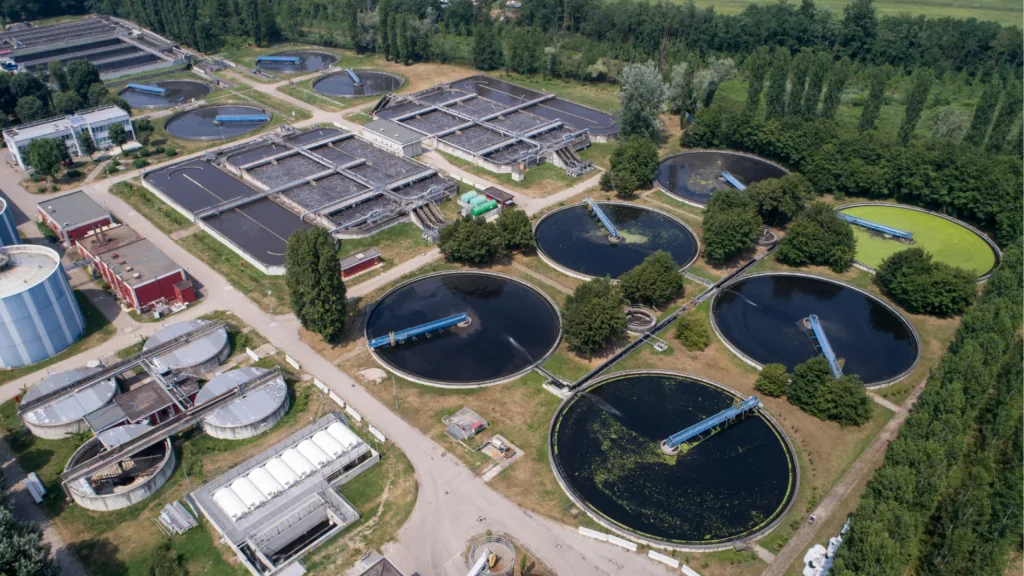 Aerial view of a wastewater treatment works, with circular clarifiers and rectangular aeration basins surrounded by trees.