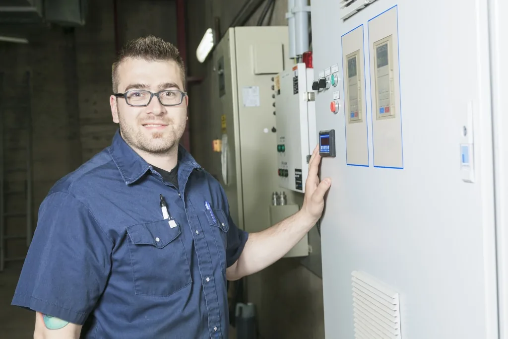 Technician in a work shirt standing beside an open electrical control cabinet, operating its panel.
