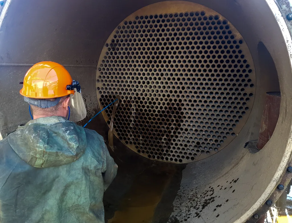 Technician in a hard hat and face shield lance-cleaning the tube sheet of an opened shell-and-tube heat exchanger, standing in water inside the shell.