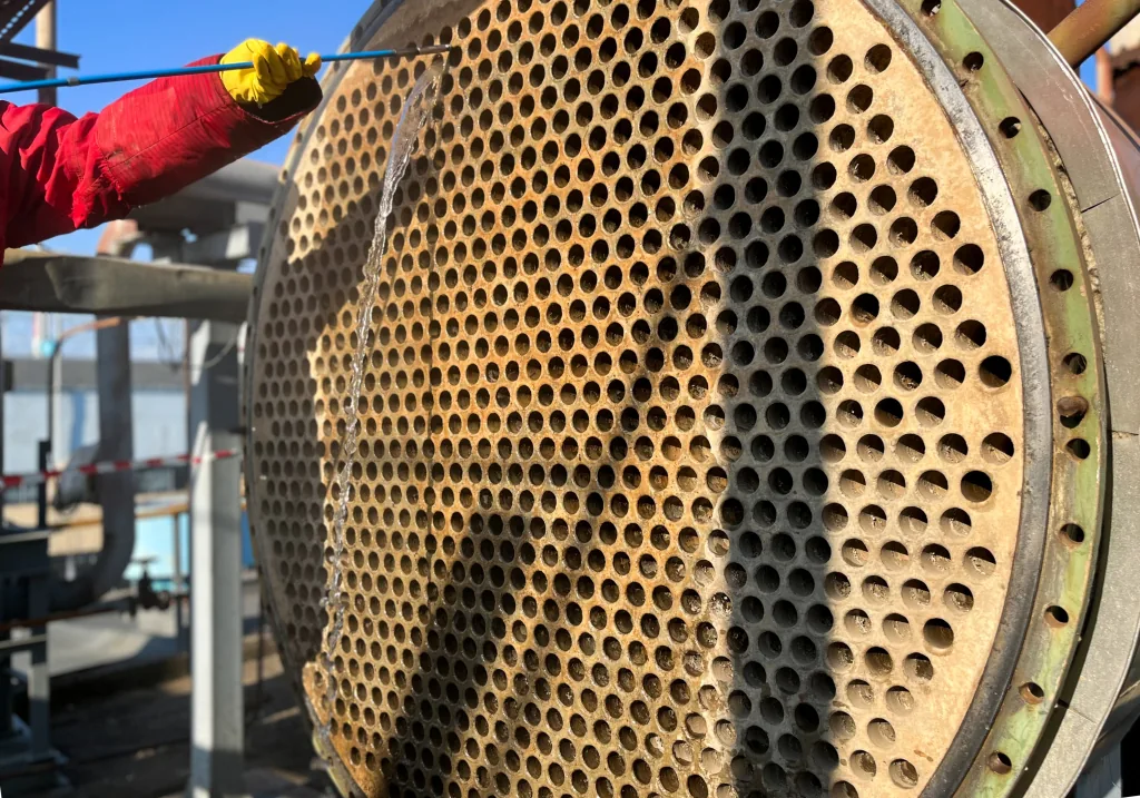 Close-up of a heavily fouled heat exchanger tube sheet outdoors, the tubes coated in pale scale, with a gloved technician cleaning one section using a lance.