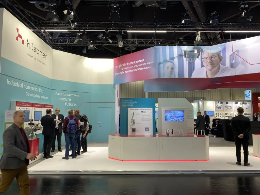 Hilscher exhibition stand at a trade fair, with industrial communication displays and visitors talking around the booth.