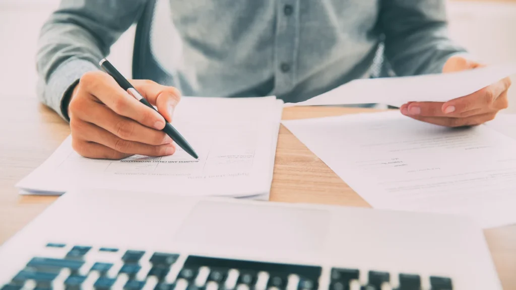 Person at a desk writing calculations on printed spreadsheets beside a keyboard.