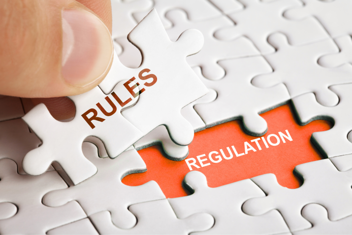 Close-up of a hand placing a white puzzle piece labeled “Rules” into a jigsaw puzzle, completing a section highlighted in orange with the word “Regulation,” symbolizing compliance and regulatory alignment.