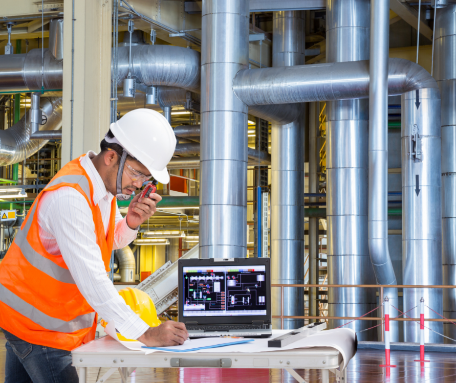 Engineer making notes on his notebook with a laptop screen on the side in a powerhouse