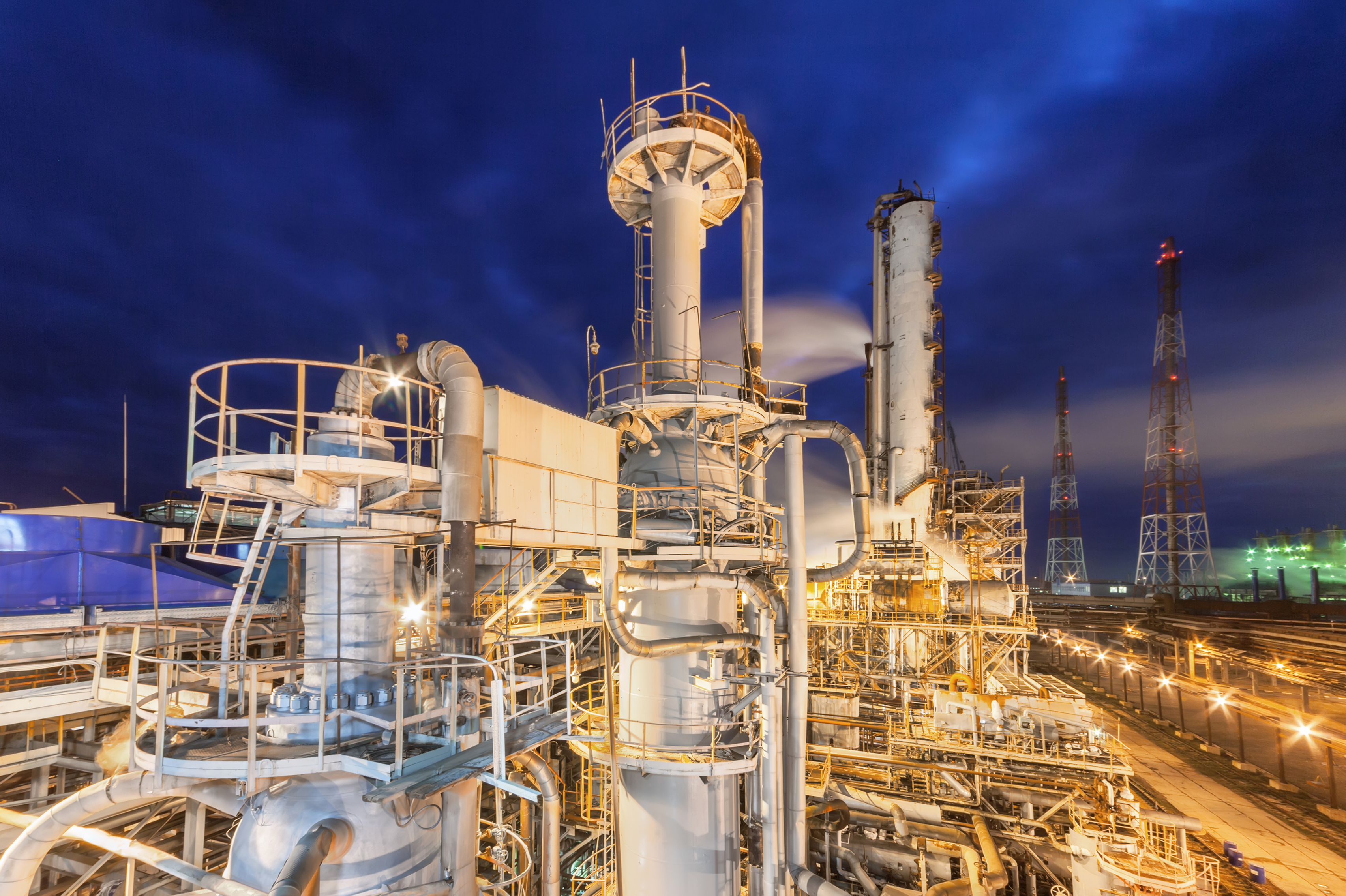 Industrial ammonia production plant at night with illuminated distillation columns, extensive piping, and refinery towers under a dark blue sky.