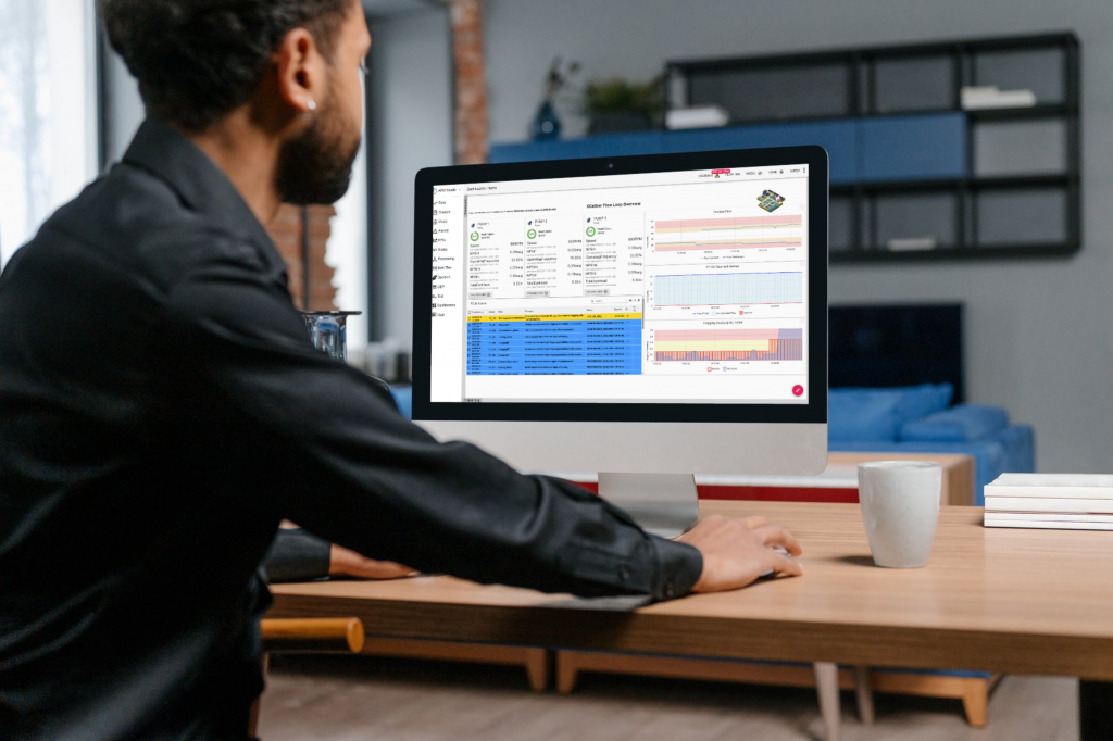 A man in a black shirt sits at a wooden desk, working on a desktop computer displaying an industrial dashboard with charts, graphs, and data tables. The background features a modern living space with shelves, a blue couch, and a coffee mug on the desk.